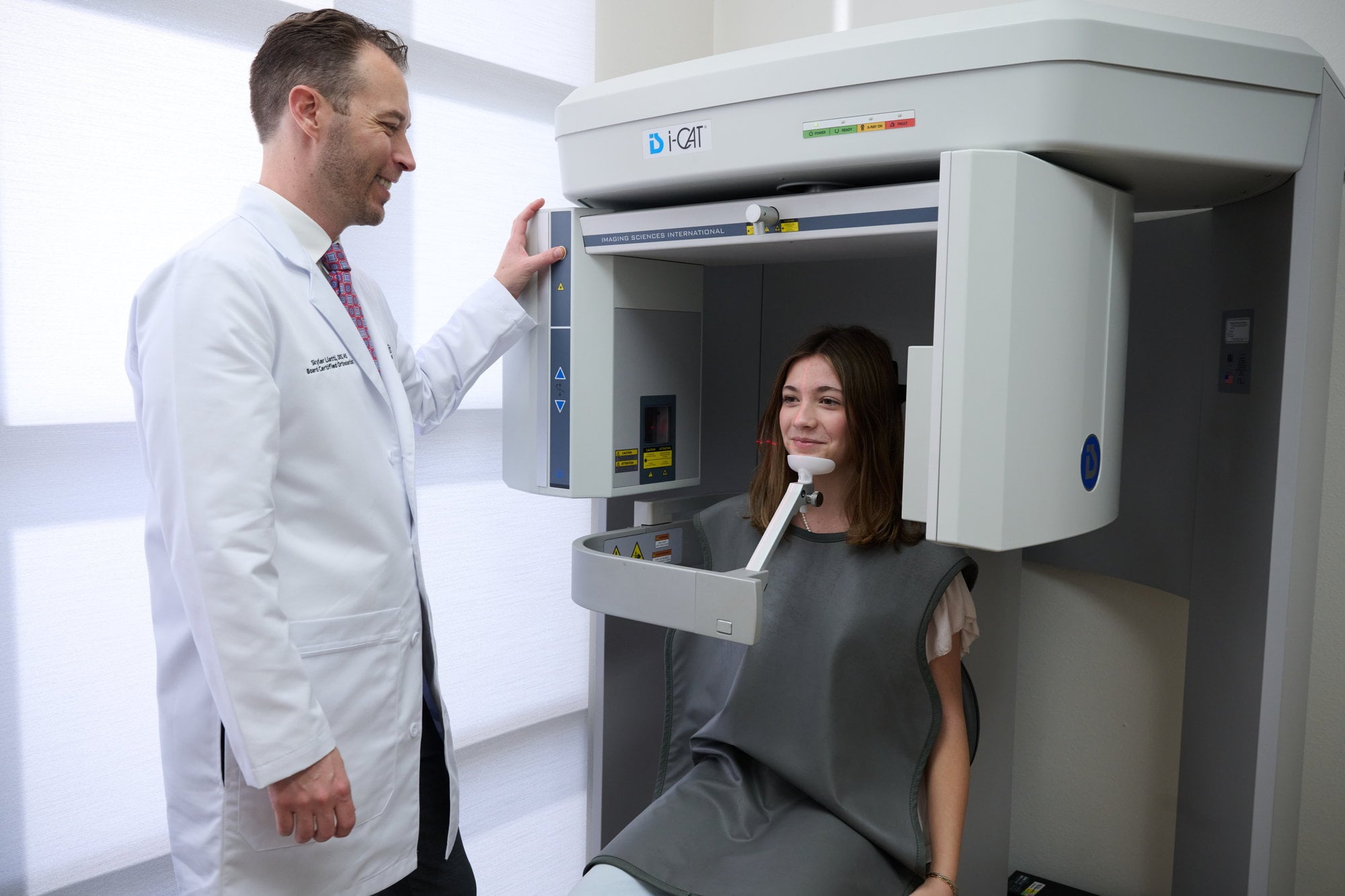 Dr. Liatti standing beside the i-CAT cone-beam CT scanner while a patient in a lead apron sits for a 3D scan at Encinitas Orthodontics